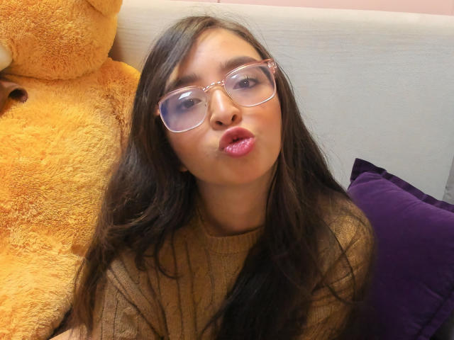 EvaMcAllister with puffy pink lips, brown wavy hair, and clear glasses, wearing a tan sweater, next to a giant yellow teddy bear and a purple pillow.