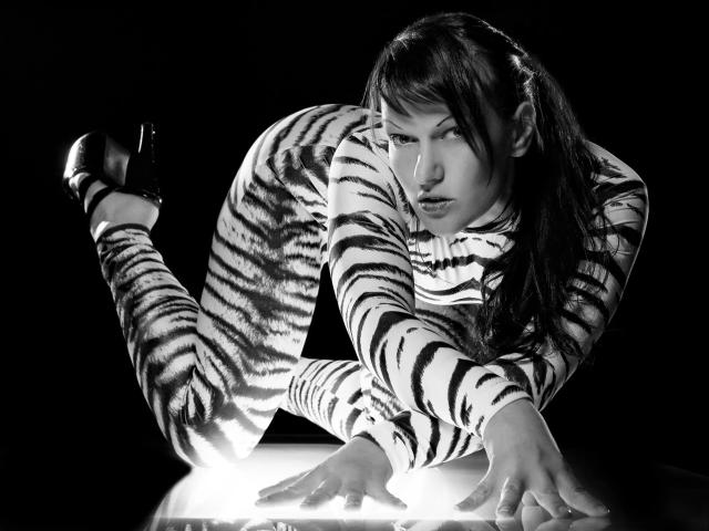 BDSM-Day is on all fours, wearing a zebra-striped outfit and black heels, with a seductive, intense look on her face, in a black-and-white, high-contrast shot.