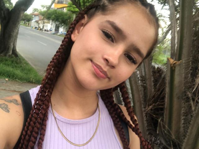 Melody Mansson ha i capelli castano intrecciati, la pelle color nocciola e piccoli tatuaggi sulla spalla. Indossa una maglietta a coste viola e sorride, con un leggero rossetto rosa..