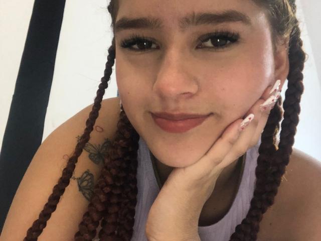 Melody Mansson, com cabelo ruivo trançado, vestindo uma regata roxa clara, com a mão apoiada na bochecha, mostrando uma tatuagem no ombro direito, e sorrindo discretamente..