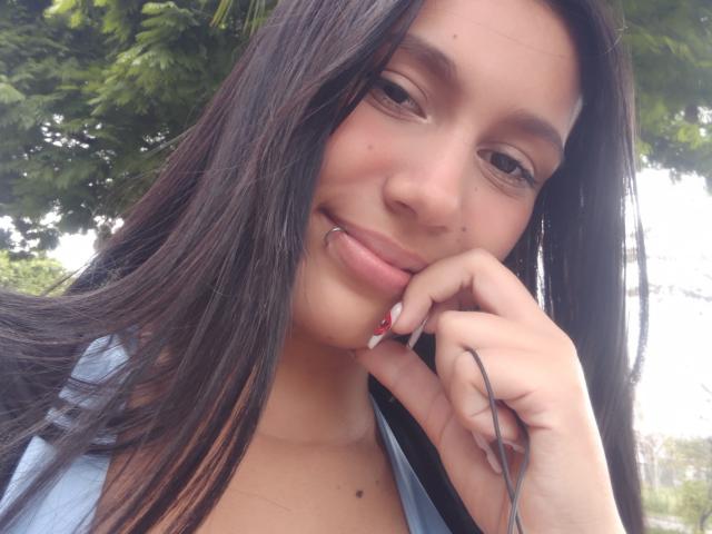 KataleyaMansson with long dark hair, light skin, brown eyes, and a slight smile, holding a phone to her cheek, showing her red-nail-painted finger and small mole on her chin.