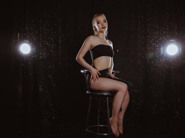 NicoleBrian mit blondierten, feuchten Haaren, die ein schwarzes Top ohne Träger und schwarze Shorts trägt, sitzt auf einem Hocker, die Beine gekreuzt, in einem dunklen, beleuchteten Studio, das mit Wasser bespritzt ist..