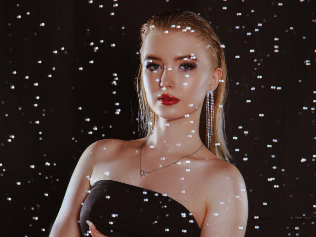 NicoleBrian con cabello rubio claro, lápiz labial rojo, vestido negro sin tirantes, aretes largos, un collar pequeño y gotas de agua brillantes en su rostro y pecho. Sus senos son visibles..