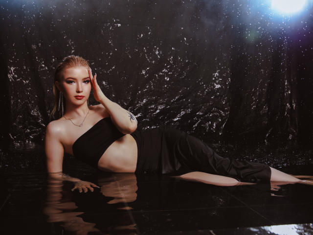 NicoleBrian with wet blonde hair, black strapless top, and black pants, lying in water, hand on head, showing tattoos, glowing light in background, looking at camera.