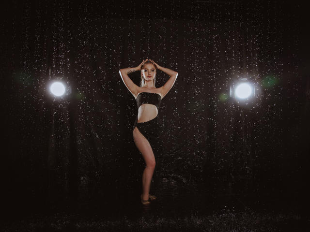 NicoleBrian stands in the rain, wearing a black, cut-out, strapless dress, arms raised, water droplets on her skin, white lights on either side, dark background.