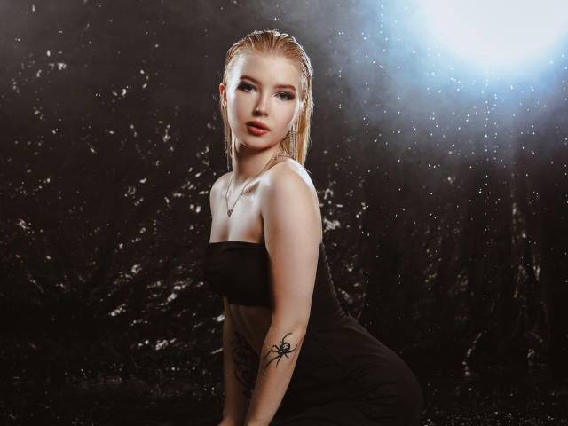 NicoleBrian, con cabello rubio claro y húmedo, vestida con un vestido negro sin tirantes, mostrando un tatuaje de araña en su brazo derecho, con agua salpicando alrededor y una fuente de luz brillante detrás..
