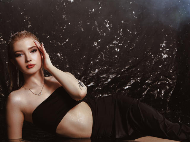 NicoleBrian with wet, blonde hair, wearing a black, strapless dress, exposing her side, hand on head, tattoo on arm, red lipstick, lying down, looking up.
