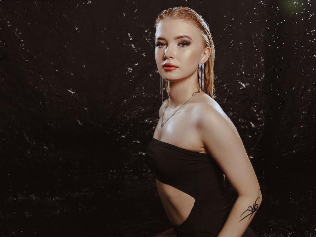 NicoleBrian, con cabello rubio claro y húmedo, vestida con un vestido negro sin tirantes, mostrando sus senos, con un tatuaje pequeño en el brazo derecho, de pie frente a un fondo oscuro con salpicaduras de agua..