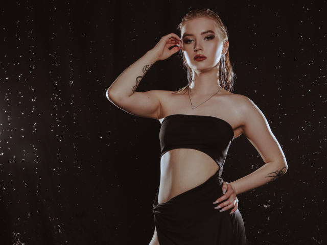 NicoleBrian with wet blonde hair, wearing a black tube top and wrap, showing tattoos on arm and hip, standing against dark background with water droplets. Boobs, slim, confident.