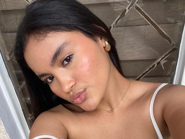 MiiaPalmer's face, showing a pout with slightly parted lips, brown eyes, and black hair. She's wearing a white top, gold necklace, and a small gold earring.