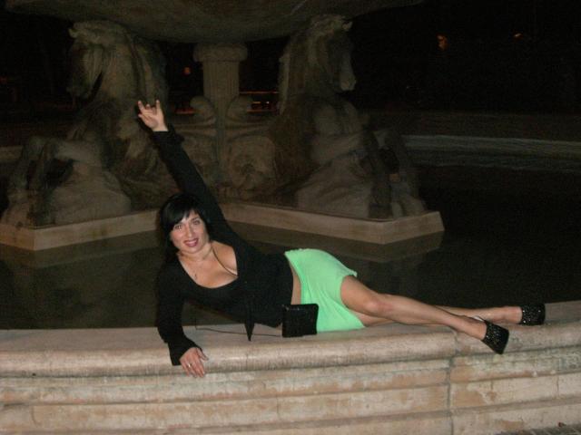Sophia01-hot with big boobs, black top, green skirt, black heels, lying on stone, raising one arm, smiling, night, water fountain, statue in background.