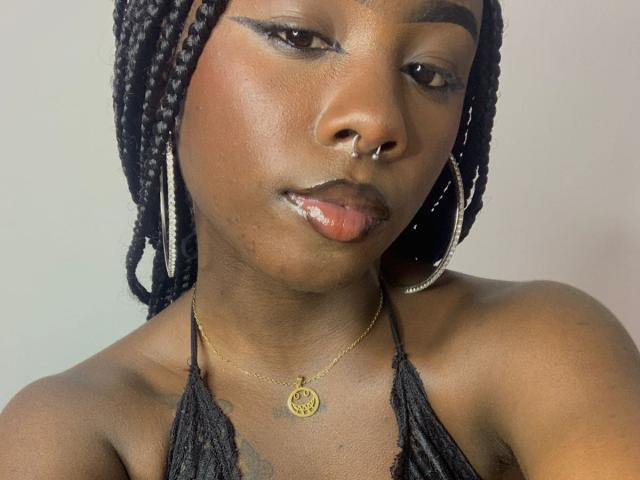 BrandyBroen has dark skin, braided black hair, a nose ring, hoop earrings, and a gold necklace with a circular pendant, showing her shiny lips and subtle makeup.