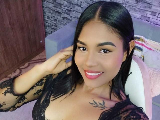 ViolettaV with black lace top, medium tits, black hair, smiling, pink lipstick, tattoo on right shoulder, hand in hair, sitting in white chair, wooden wall in background.