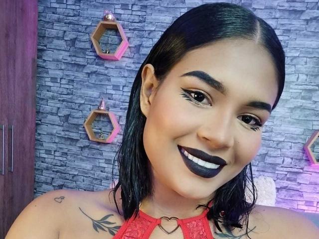 ViolettaV has black lipstick, medium brown skin, straight black hair, and a heart tattoo near her right shoulder. She's smiling with her tits partially visible in a red lace top.