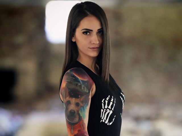 Jane Warren, avec de longs cheveux bruns, un débardeur noir laissant apparaître un motif osseux blanc, un tatouage de chien coloré sur le bras droit, regardant l'appareil photo avec un léger sourire. Arrière-plan flou..