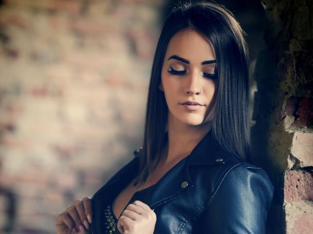Jane Warren, met lang bruin haar, draagt een zwarte leren jas, kijkt naar beneden met gesloten ogen, en laat haar aantrekkelijke eyeliner en subtiele make-up zien, terwijl ze tegen een bakstenen muur staat..
