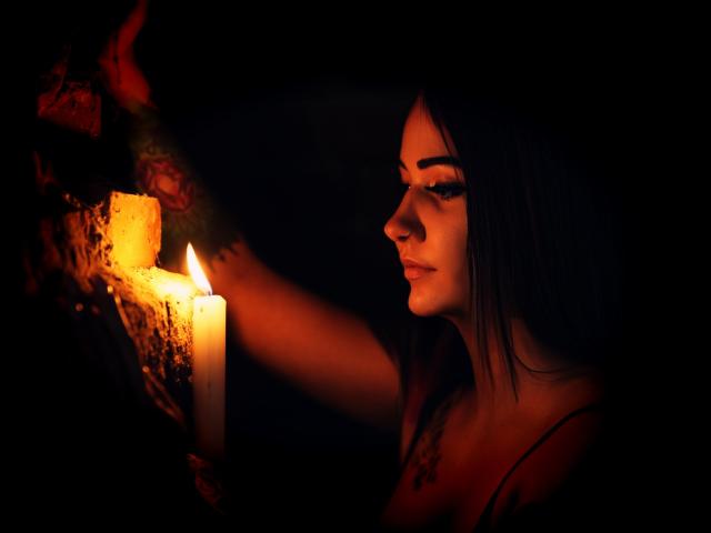 JaneWarren with dark hair, looking at a lit candle, holding a rough stone. Candle's flame casts warm orange light on her face, highlighting her eyes and nose.