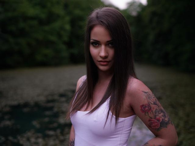 Jane Warren, con cabello castaño largo, camiseta blanca, brazo tatuado y expresión seria, está de pie junto a una vía fluvial verde y boscosa, mostrando sus senos y un pequeño tatuaje en la cadera..