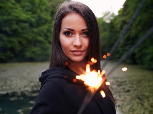 Jane Warren mit langem, braunen Haar, grünen Augen und einer kleinen Nase, die ein funkelndes Feuerwerk hält, trägt eine schwarze Kapuzenjacke, lächelt leicht und befindet sich in einem grünen Wald..