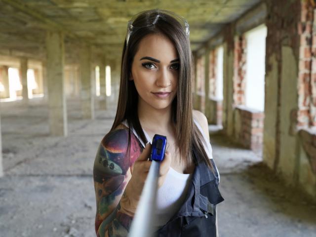 Jane Warren, con cabello castaño largo, un brazo tatuado y una blusa blanca, sosteniendo una cámara azul, tomándose un selfie en una habitación deteriorada, iluminada por el sol y con paredes de ladrillo. Sus senos son visibles..