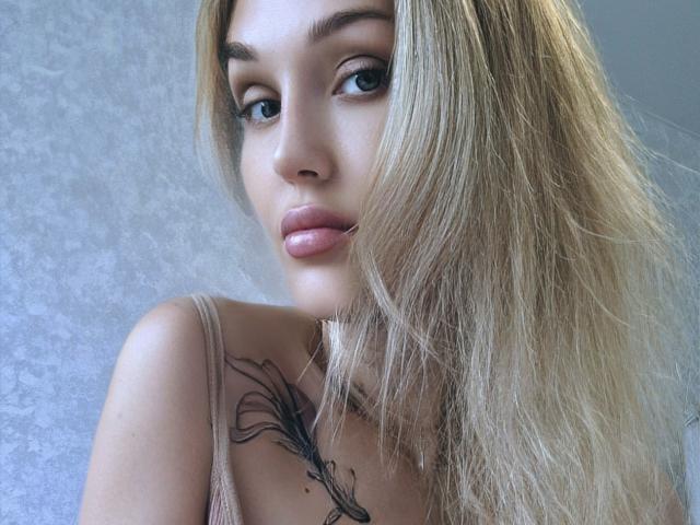 Angelovna with blonde hair, blue eyes, and pink lips, showing a tattoo on her chest, wearing a beige top, looking at the camera with a slight pout.