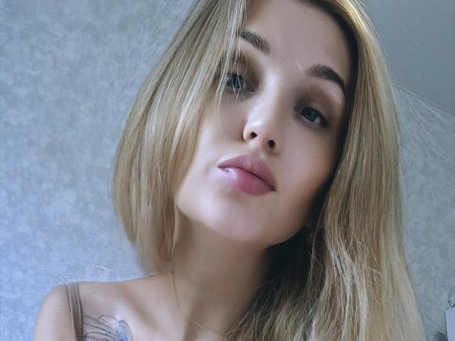 Angelovna with blonde hair, full pink lips, and a tattoo on her left chest, staring at the camera with a slight, confident smile. Her eyebrows are dark and well-groomed.