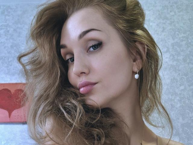 Angelovna has wavy brown hair, blue eyes, and pink glossy lips. She's wearing a diamond earring and has a cute, confident expression. Her tits are subtly visible.