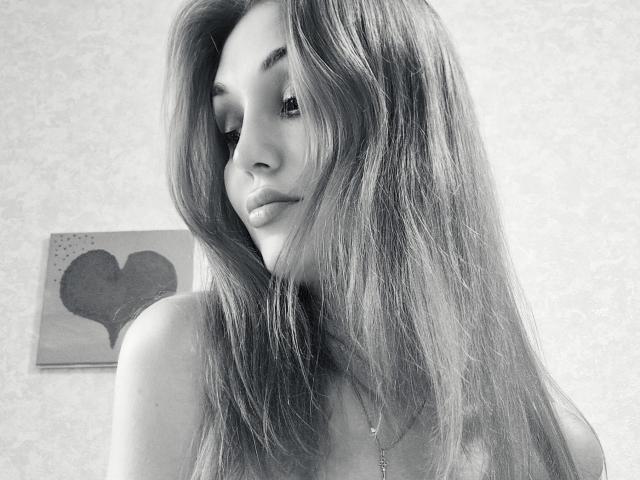 Angelovna with long, straight, dark hair covering her right eye, looking slightly downward with a subtle smile, wearing a necklace, in a black-and-white photo.