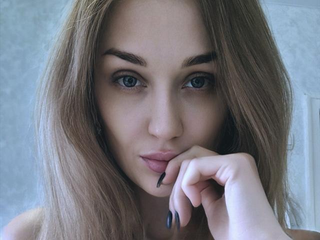 Angelovna with light brown hair, blue eyes, and black nails, biting her finger with a subtle, sexy smile, showing off her smooth, pale skin and full lips.