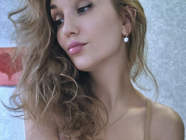 Angelovna with wavy blonde hair, pink glossy lips, subtle eyeliner, diamond earrings, and a tan strap top, showing off her smooth, fair skin and cute, feminine face.