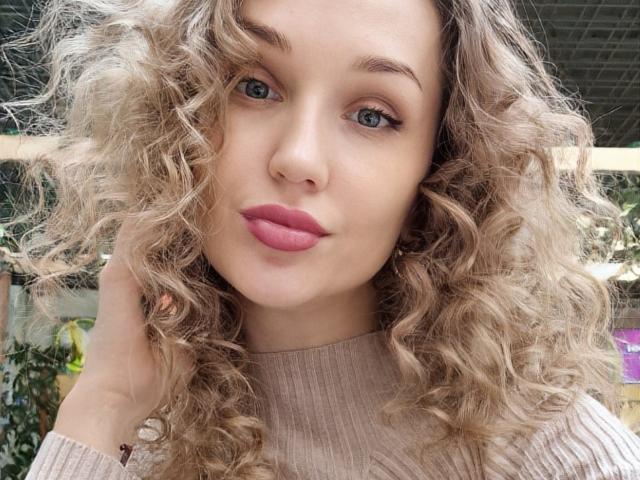 Angelovna with curly blonde hair, pink lipstick, and blue eyes, wearing a beige ribbed shirt, hand in hair, looking at camera. She's got a cute, sexy face.