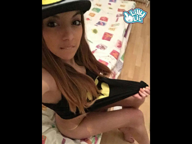 Lilly-Lil-hot with long brown hair, wearing a black shirt with a yellow logo, lifting it to reveal her bare ass, sitting on a bed with colorful patterns.