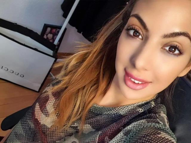 Lilly-Lil-hot with blonde hair, brown eyes, and pink lipstick, smiling at the camera, wearing a camouflage-patterned shirt, in a selfie. Her tits are not visible.