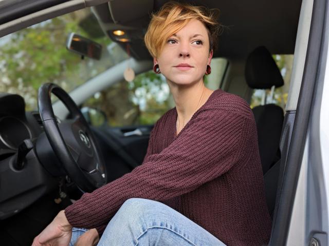 MiaSonne-hot sitting in a car, wearing a maroon knitted sweater, blue jeans, and red earrings, with short, messy blonde hair, looking forward with a serious expression.