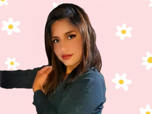 AriGame with medium brown hair, dark green top, hand on shoulder, subtle smile, pink background with white daisy patterns, makeup includes winged eyeliner and red lipstick.