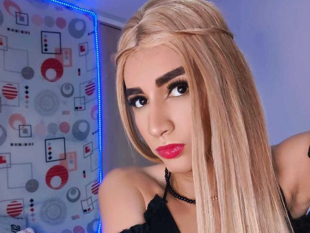 BritS hat langes, blondes Haar, dicke Augenbrauen, roten Lippenstift und eine schwarze Halskette. Sie blickt zur Seite und hat einen ernsten Gesichtsausdruck. Ihre Brüste sind teilweise sichtbar..