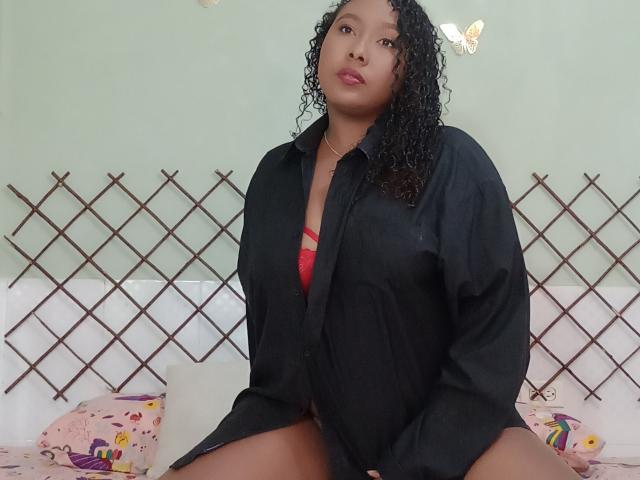 CaroldelaTorre with curly black hair, wearing an open black shirt, exposing red bra, sitting with legs apart, looking at camera, green wall, butterfly decals, on bed with colorful pillows.