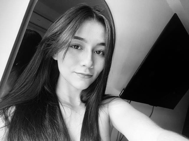 DeisyWest has long dark hair, light skin, and a slight smile. She's topless with small tits, looking at the camera. Black and white selfie.