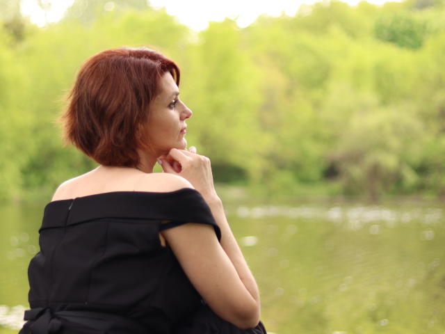 LisaTreds with red hair, black off-shoulder top, looking sideways, hand near mouth, standing by a green, blurry water background. Her tits are slightly visible from the side.