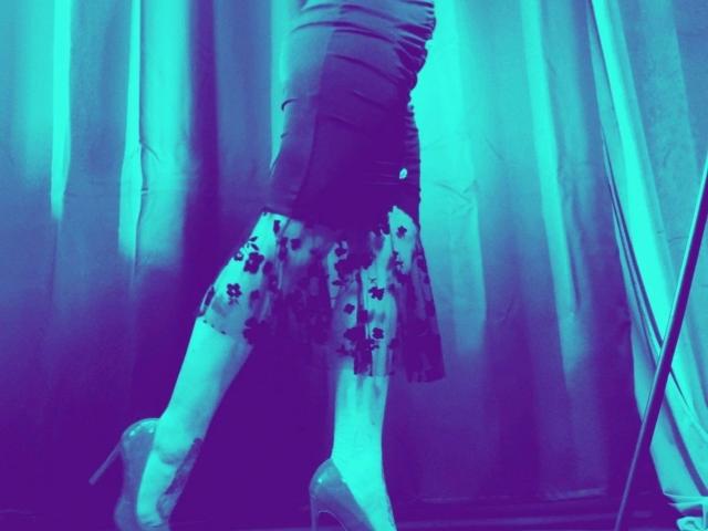 ScorpionLady-hot's legs, wearing a ruffled skirt with black floral patterns, white stockings, and clear high heels, under blue neon lighting.