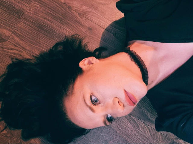 ScorpionLady-hot lies on wooden floor, head upside down, black curly hair, pale skin, black choker, blue eyes, black top, looking up with slightly parted lips, soft expression.