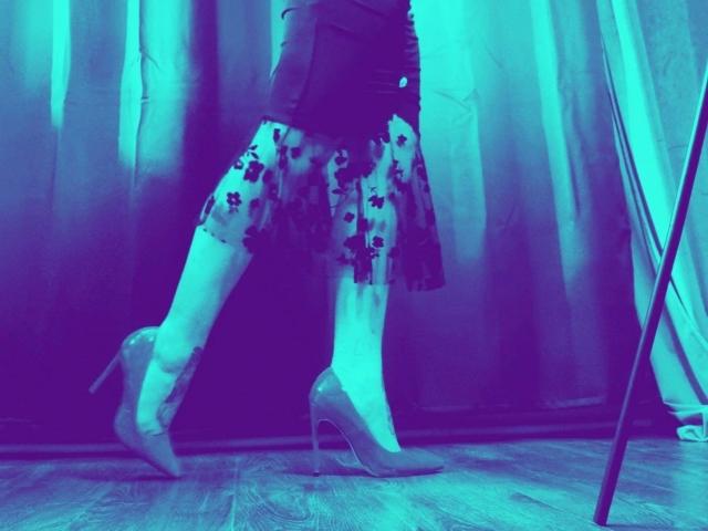 ScorpionLady-hot's legs wearing a black top and floral skirt with transparent high heels, standing on wooden floor, blue-tinted light, her ass slightly visible under skirt.