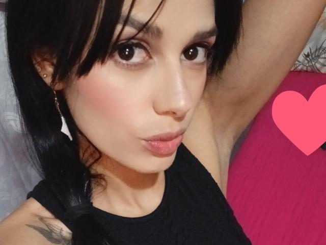 Anais Hilton mit schwarzen Haaren, braunen Augen, rosiger Rouge und vollen Lippen, trägt ein schwarzes Oberteil, ein Arm ist erhoben, über ihrer nackten Brust befindet sich ein pinkes Herz, sie hat ein kleines Tattoo an ihrer Schulter..