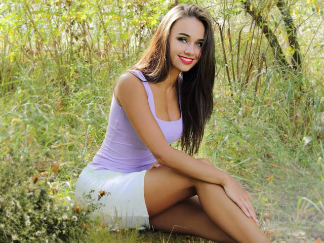 ManuelaRose, con el pelo largo y castaño, vistiendo una camiseta morada y una falda blanca, sentada con las rodillas levantadas, sonriendo, mostrando sus pechos y glúteos, en un entorno natural al aire libre..