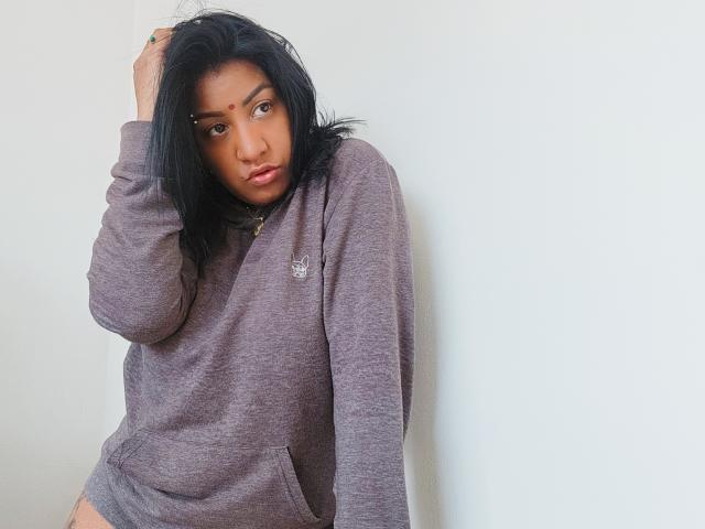 Nahirr with black hair, wearing a purple hoodie, hand in hair, looking serious, soft brown skin, slight pout, subtle makeup, white wall background, casual style, relaxed pose.