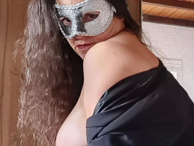 Lady-Mercedes with long wavy brown hair, wearing a glittery silver mask, black off-shoulder top, and looking over her shoulder, showing her bare tits.