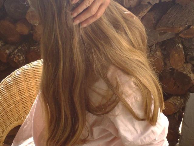 Reallive1-hot's long blonde hair, held by a hand, covering her pink shirt. She's sitting on a wicker chair, with a stack of logs in the background.