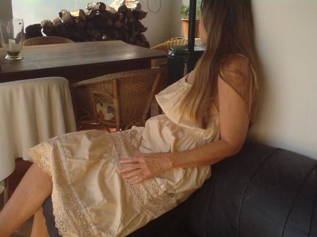 reallive1-hot is sitting on a black couch, wearing a beige lace dress, with long brown hair, showing her ass and hands on her lap.