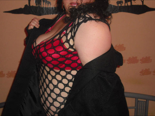 REALAMATEUR-hot with curly black hair, wearing a red and black fishnet top, exposing her tits, and black coat falling off her shoulders. Her skin is pale.