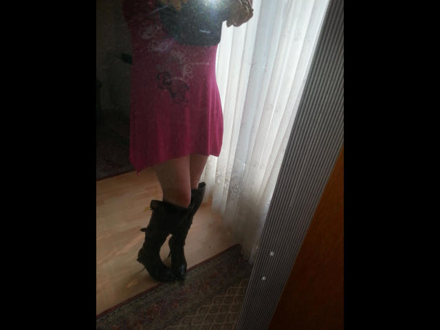 Mirror selfie of monelive-hot in a pink dress with butterfly design, standing in black boots, holding camera, white curtains, wooden floor, and patterned rug in background.